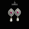 A pair of ornate silver-toned drop earrings with bright pink teardrop-shaped center stones and dangling white pearl drops, displayed on a black stand against a gray background. The brand name 'SAHELI MY BEST FRIEND' is visible at the top.