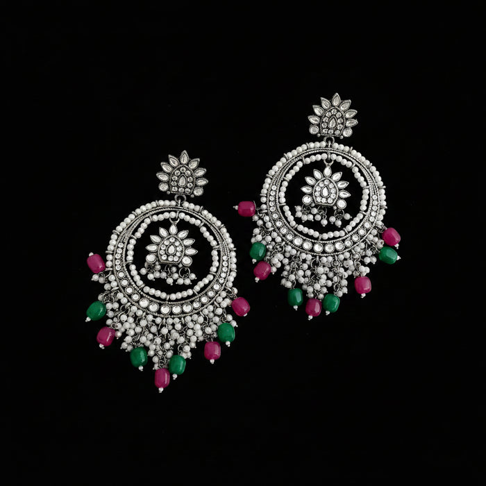 A pair of ornate silver-toned dangle earrings with intricate beadwork and dangling pink and green gemstone accents, displayed on a dark reflective surface.