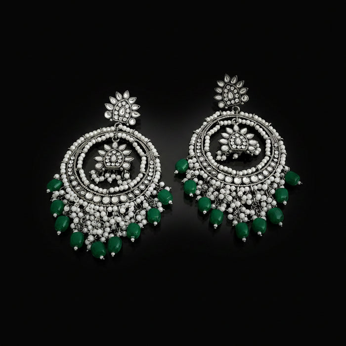 A pair of silver-toned Indian chandelier earrings decorated with white beads, clear stones, and green bead accents, displayed on a dark reflective surface.