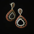 A pair of ornate teardrop-shaped earrings with a black center, red and gold floral patterns, and a silver-tone border set with clear stones, displayed on a dark reflective surface.