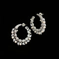 A pair of silver-toned crescent-shaped hoop earrings decorated with leaf and pearl motifs, displayed on a dark reflective surface.