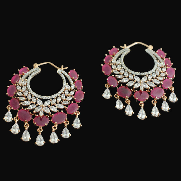 A pair of gold-plated hoop earrings decorated with pinkish-red round gemstones, clear pavé-style stones, and dangling clear teardrop-shaped crystals, displayed on a dark reflective surface.