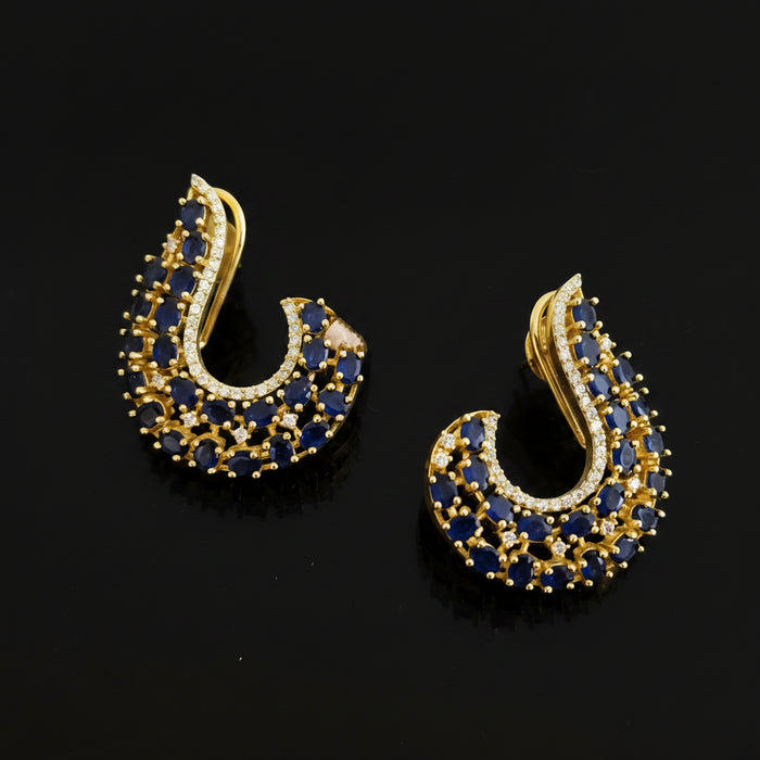 A pair of gold crescent-shaped stud earrings embellished with deep blue round stones and small clear accent stones, displayed on a dark reflective surface with the SAHELL MY BEST FRIEND logo above.