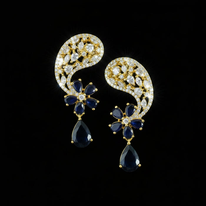 A pair of gold-toned traditional Indian jhumka earrings with deep blue stones and clear pavé accents, displayed on a dark reflective surface with a Saheli by Srinivasand logo in the top left corner.