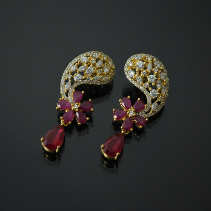 A pair of gold crescent-shaped earrings decorated with clear crystals and red teardrop gemstones, displayed on a black reflective surface with a SAHELI MY BEST FRIEND logo in the top right.