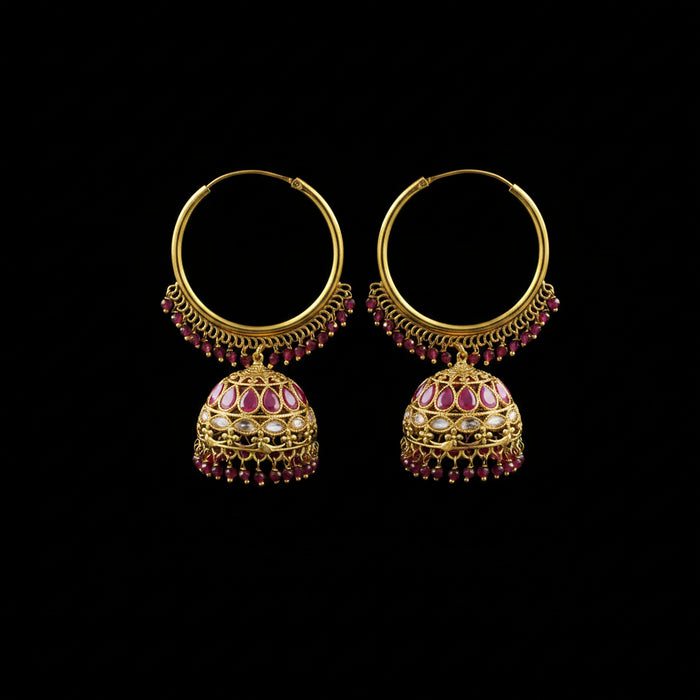 A pair of ornate gold-toned hoop earrings with dangling jhumka-style bells, decorated with pink gemstones and small red bead drops, displayed on a black stand against a gray background.