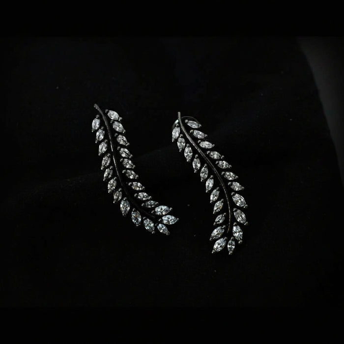A pair of leaf-shaped drop earrings with marquise-cut clear stones set in a dark metal, displayed on a black velvet surface with the SAHELI MY BEST FRIEND logo in the top right.
