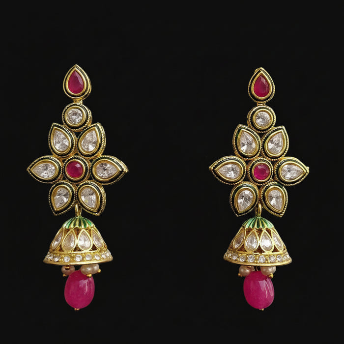 A pair of gold-plated Indian jhumka earrings with pink stone accents, clear crystals, and green enamel details, displayed on a black Y-shaped stand against a gray background. The brand logo 'SAHELI MY BEST FRIEND' is visible at the top.