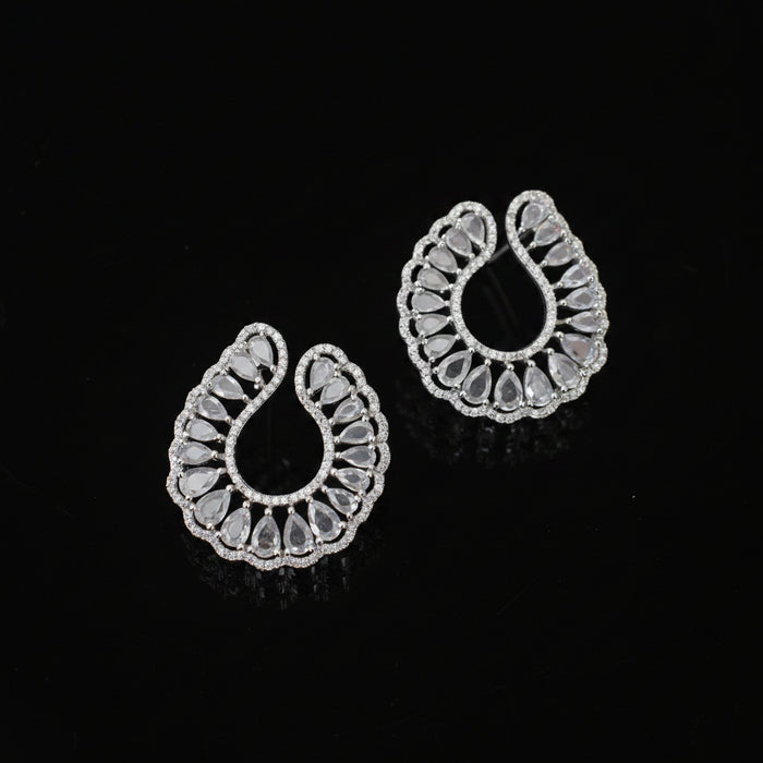 A pair of silver-toned teardrop-shaped earrings with intricate filigree cutouts and clear stones, displayed on a dark reflective surface.
