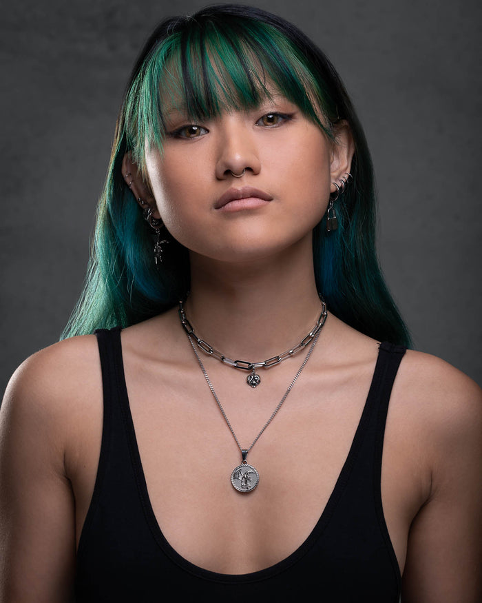 A young woman with long green hair and bangs stares confidently at the camera. She wears a black tank top, layered silver necklaces including the Fallen Angel Medallion Pendant, and multiple earrings. The background is dark and plain.