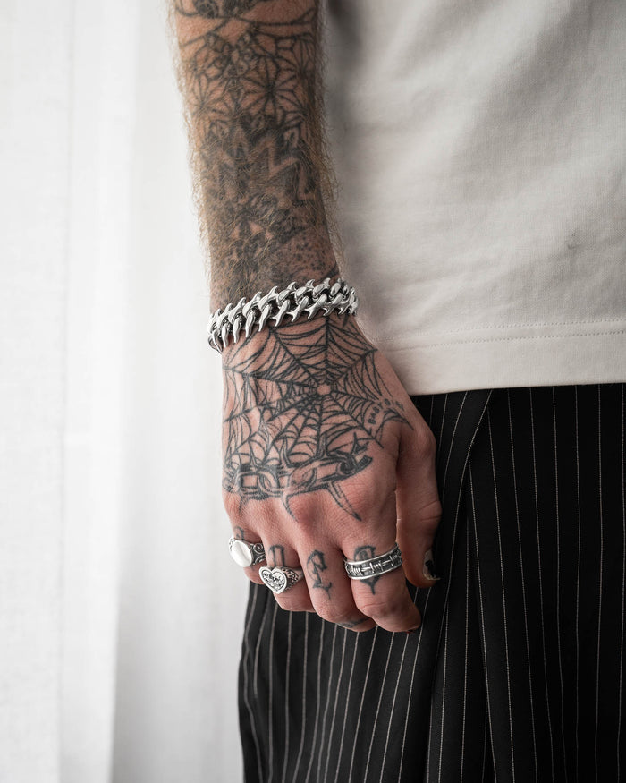 A tattooed hand adorned with silver rings and the bold "Fishbone" Spiked Cuban Bracelet in 316L stainless steel rests by their side. Dressed in a white shirt and black pinstriped pants, they showcase a prominent spiderweb tattoo design.