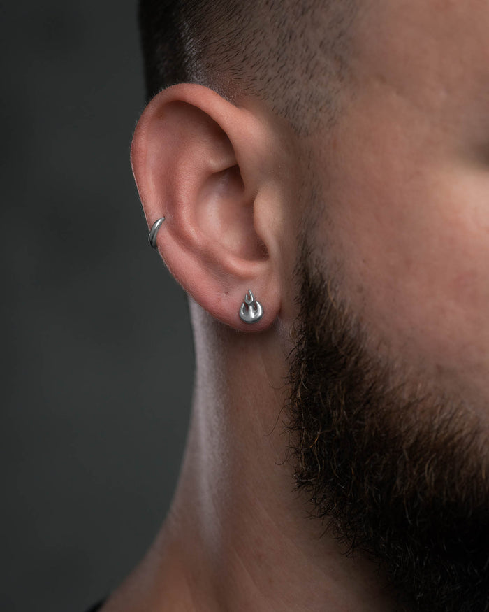 Close-up of a person with short hair and a beard wearing the Flame Stud Earring in the earlobe. The stainless steel design stands out against a dark, blurred background.
