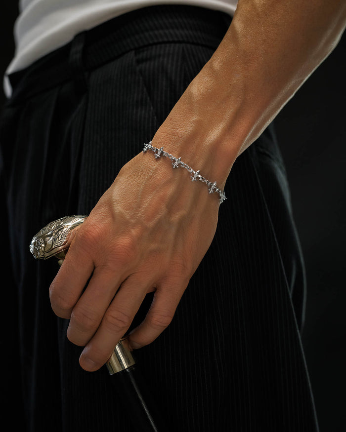 A close-up of a person wearing the Statement Collective "Fleur De Lis" 10mm Bracelet, holding an ornate cane. The person is dressed in black pinstripe pants and a white shirt, set against a black background.