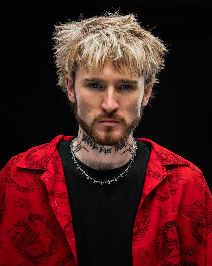 A person with shaggy blond hair, a full beard, and neck tattoos wears Statement Collective’s "Fleur De Lis" 10mm Chain along with a red patterned shirt layered over a black top, staring seriously at the camera against a dark background.