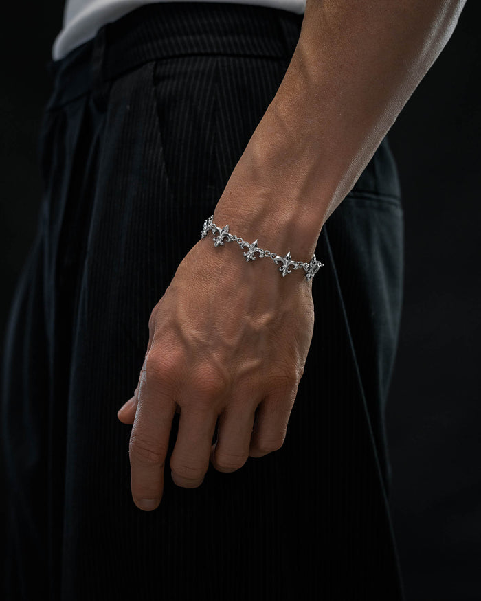 A close-up of a person's hand wearing the "Fleur De Lis" 15mm Bracelet by Statement Collective, featuring intricate silver star-shaped links. The person is dressed in black pinstriped pants and a white shirt, set against a dark background.