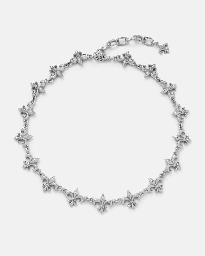 The "Fleur De Lis" 15mm Chain by Statement Collective features linked fleur-de-lis charms evenly spaced along a silver chain, with an adjustable clasp and a dangling charm at the end, displayed on a plain white background.