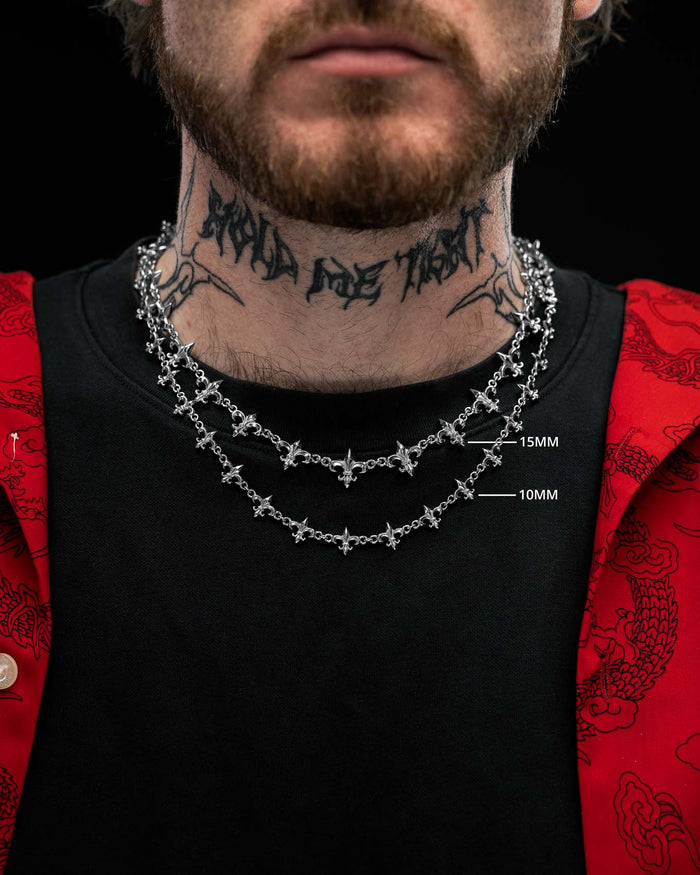 Close-up of a person wearing two silver cross necklaces of different thicknesses, including the “Fleur De Lis” 10mm Chain by Statement Collective, with a tattooed neck, black shirt, and a red jacket featuring dragon patterns.