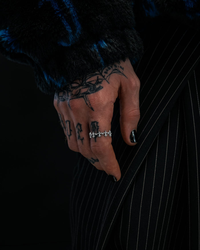 A tattooed hand with black nail polish wears the “Fleur De Lis” Ring by Statement Collective, a silver, stackable piece featuring star-shaped details, resting on pinstriped pants. The person also wears a dark, furry textured jacket.