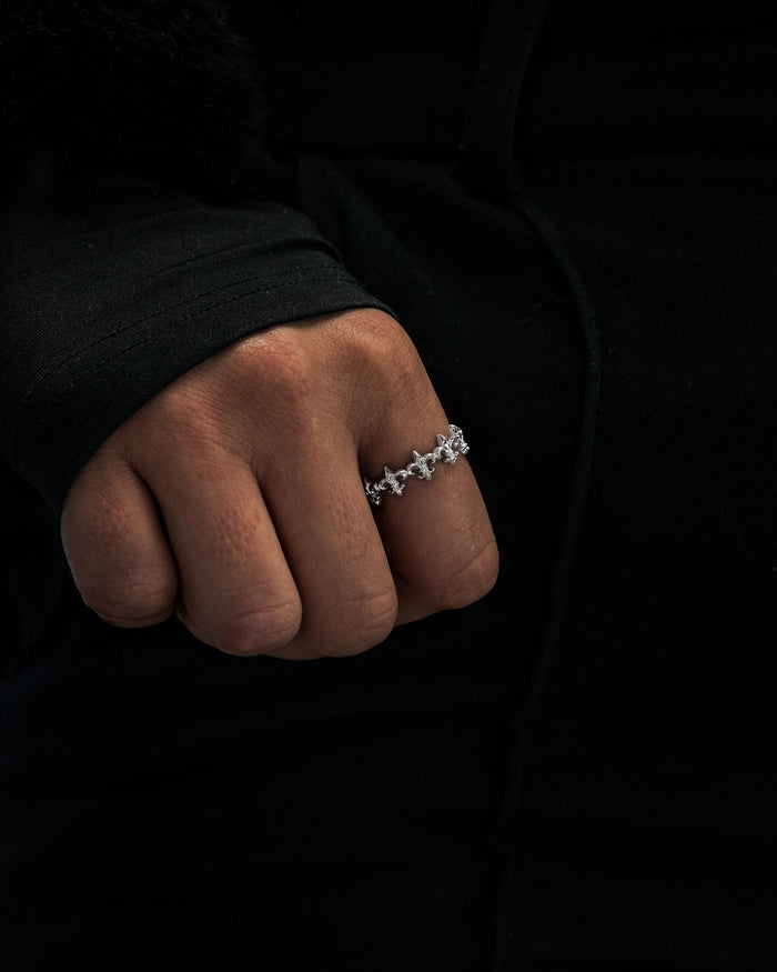 A hand adorned with Statement Collective’s “Fleur De Lis” ring, featuring a silver floral pattern, rests against a dark, long-sleeved garment.