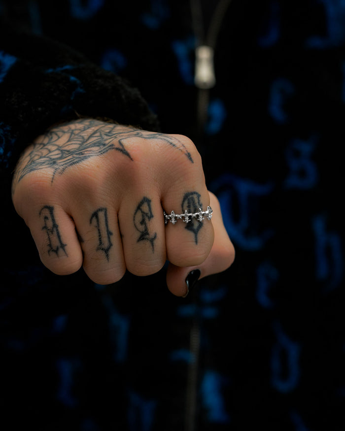 A close-up of a tattooed hand with a spiderweb design and bold letters on the fingers, adorned with Statement Collective’s striking “Fleur De Lis” silver ring alongside a sleek stainless steel band. The background features dark fabric with blue patterns.