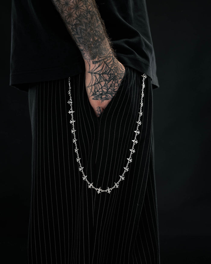 A person with a spiderweb tattoo on their hand wears black pinstripe pants, a black shirt, and the ornate “Fleur De Lis” Wallet Chain by Statement Collective hanging from their pocket, set against a dark background.