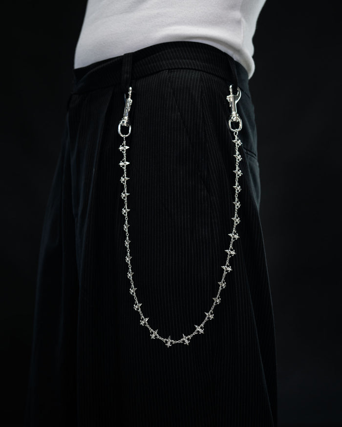 A person wearing black pinstripe pants with the “Fleur De Lis” Wallet Chain by Statement Collective, crafted from decorative sterling silver and attached at the waistband, standing against a dark background.