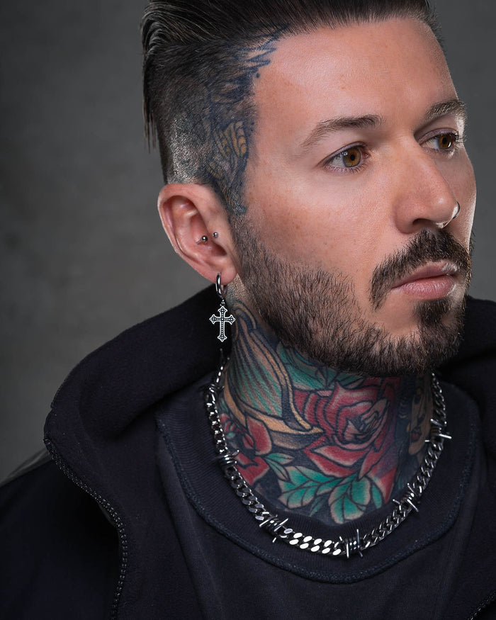 A man with slicked-back dark hair, facial hair, and neck tattoos wears a French Cross Earring, a spiked chain necklace, and a black jacket against a neutral background.