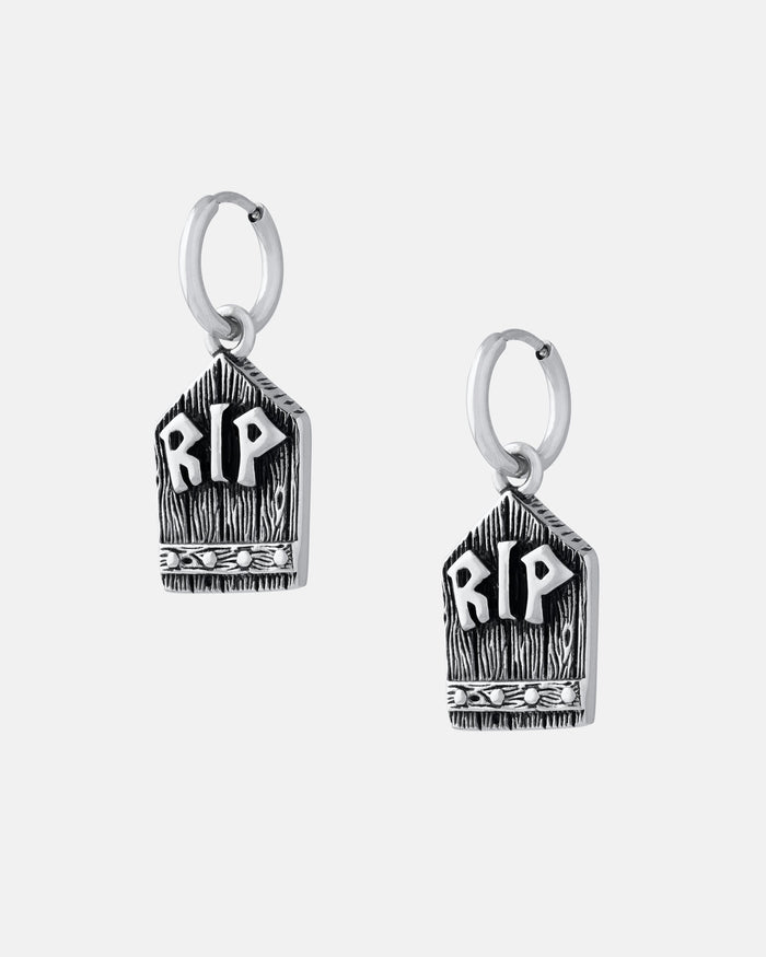 The Gravestone Earring is a silver hoop earring with tombstone-shaped pendants engraved with RIP and decorative details, offering a witchy style—shown on a plain light gray background.