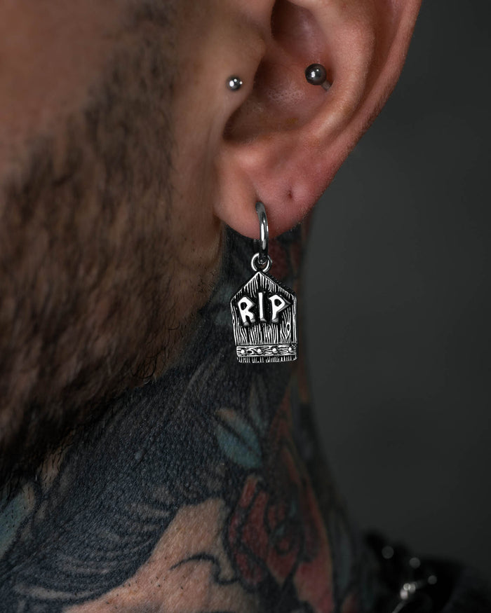 A close-up of a person's ear with two piercings, one wearing the Gravestone Earring engraved with RIP. Colorful tattoos on their neck enhance the unique and witchy vibe of this standout jewelry piece.
