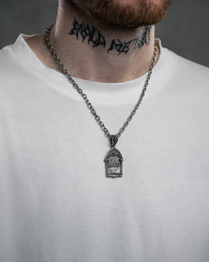 A person in a plain white shirt shows a neck tattoo with stylized black lettering and wears the Gravestone Pendant, a silver necklace with intricate design, blending subtle style and macabre jewelry. The lower face and neck are visible.