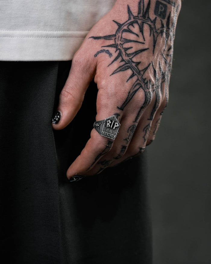 A tattooed hand with black nail polish and white dots wears the Gravestone Ring, a bold piece for lovers of graveyard-themed jewellery. The large ring, marked "RP," stands out against a dark garment and intricate tattoos.