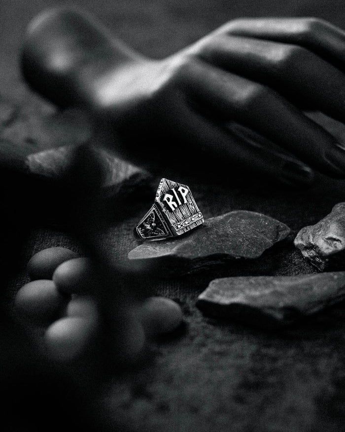 A black and white photo shows a hand near scattered stones and round objects, featuring the Gravestone Ring—a macabre, tombstone-shaped accessory engraved with RIP—placed among the rocks.