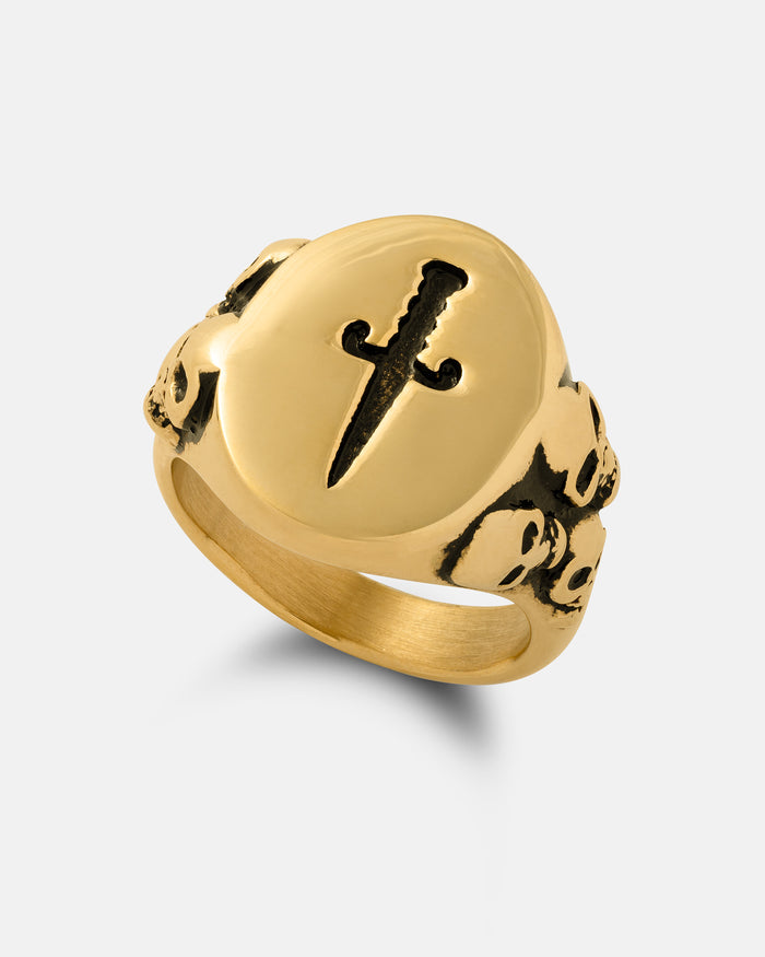 The Graveyard Signet Ring (Gold) by Statement Collective showcases a raised dagger motif on the front, complemented by intricately engraved skulls along the band. Set against a simple light background, this striking piece offers bold gothic symbolism paired with timeless elegance.