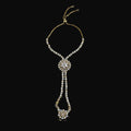 Gold-toned chain necklace with multiple rows of clear rhinestones, featuring a central oval cluster and a dangling floral pendant, displayed on a solid pink background.