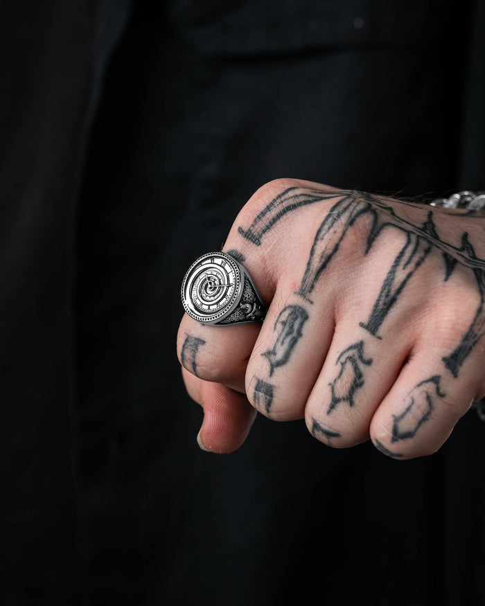 A close-up shows a tattooed hand wearing the Infinite Clock Ring, a large, ornate stainless steel piece with an intricate circular design. The wearer is dressed in black, and the tattoos have bold, dark lines and shapes.