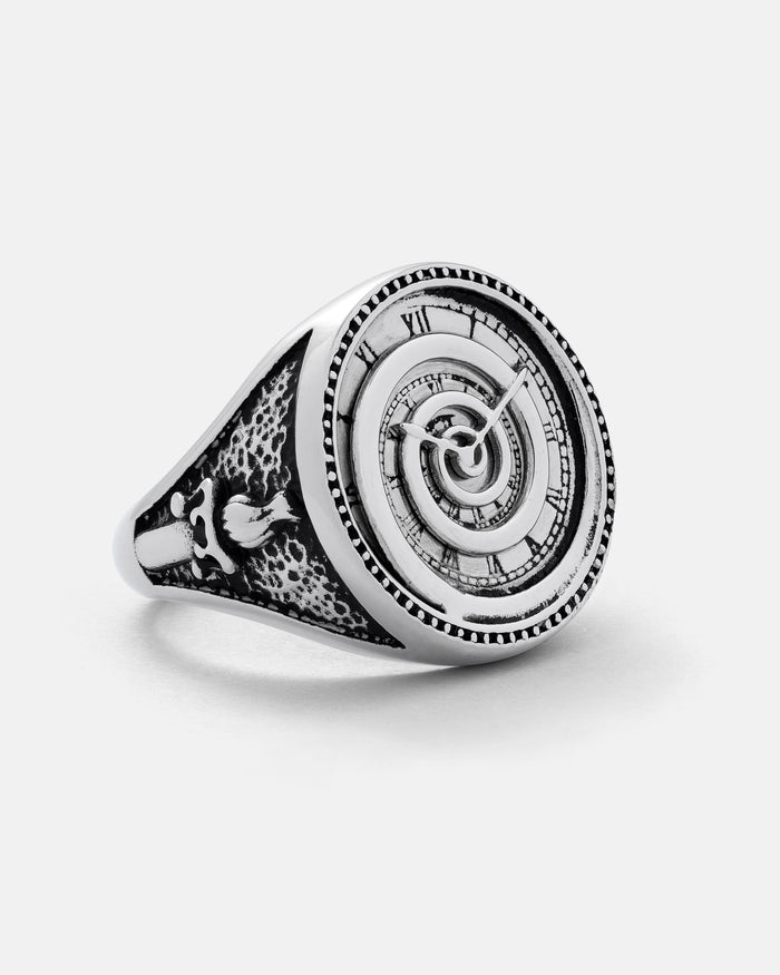 Introducing the Infinite Clock Ring by Statement Collective: a silver ring showcasing an intricately engraved spiral clock face with Roman numerals, complemented by detailed band patterns and a distinctive bird motif on the side.