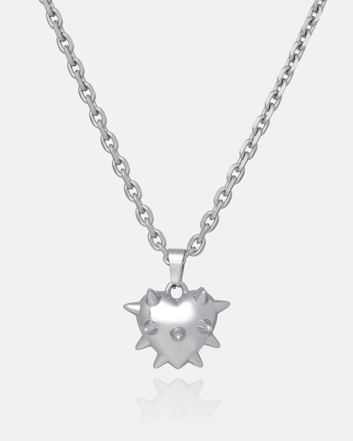 The "Love Hurts" Spiked Heart Pendant is a silver necklace with a shiny heart-shaped pendant, raised spike details, and a central gemstone, shown on a plain white background for an edgy grunge look.