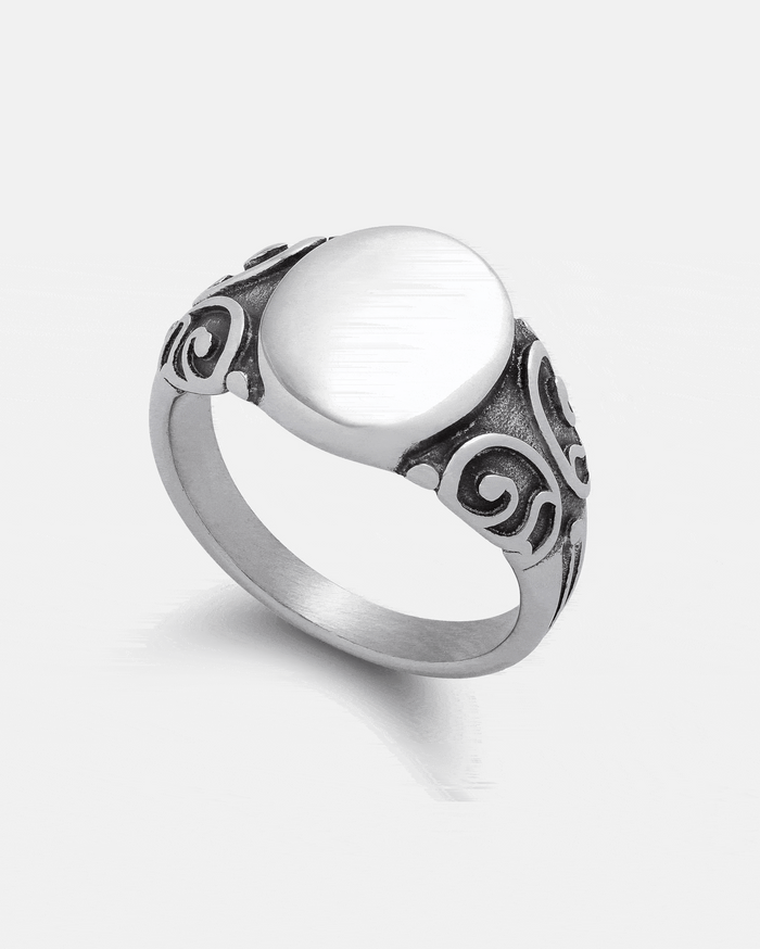 The Statement Collective Mini Oval Scroll Signet Ring features a plain, miniature oval signet centerpiece crafted from 316L stainless steel, with ornate traditional scrollwork engraving on both sides of the band, shown against a white background.