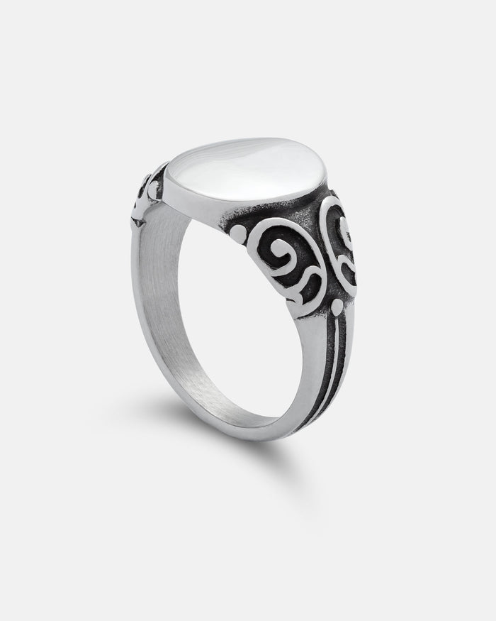 The Mini Oval Scroll Signet Ring by Statement Collective is a silver ring featuring a smooth white stone atop an oval setting, with black traditional scroll pattern designs adorning the band, displayed against a plain white background.