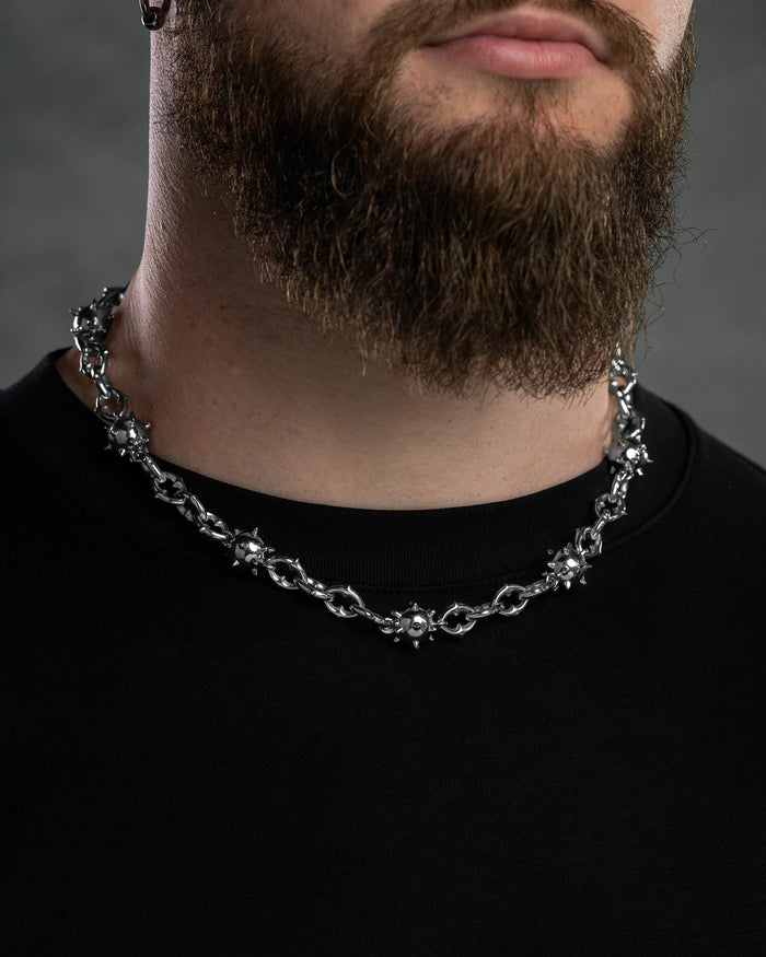 A close-up of a man with a thick brown beard wearing the "Morning Star" Cathedral Chain, a chunky stainless steel necklace with spiked ball-shaped details, and a black crew-neck shirt against an out-of-focus neutral background.