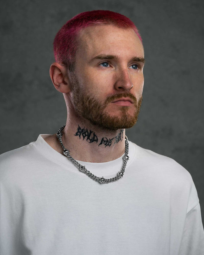 A person with short pink hair, a beard, blue eyes, and a neck tattoo wears a white shirt and the "Morning Star" Cuban Chain. The background is plain dark gray.