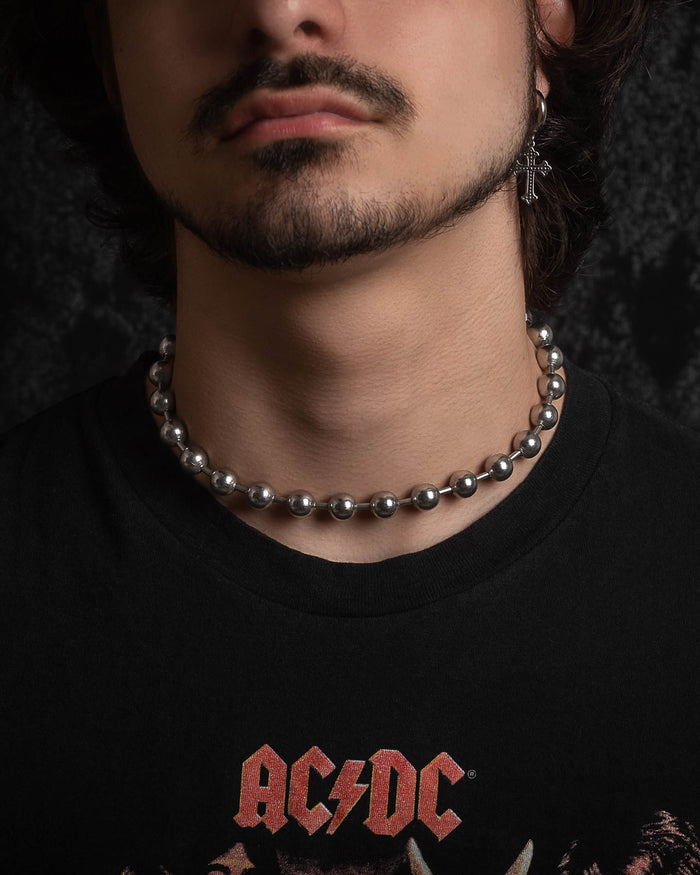 A person with a short beard and mustache wears the No Tourists Choker, a cross earring, and a black AC/DC T-shirt against a dark background.