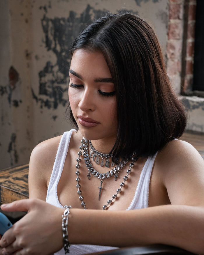 A young woman with straight dark hair and layered silver necklaces from the Crybaby Set by Statement Collective, including a delicate small neck choker, looks down at her phone while sitting indoors at a rustic wooden table. Peeling paint and exposed brick are visible in the background.
