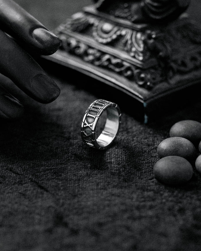 A black and white close-up of the Numerals Ring on a textured surface, with a reaching hand, round stones nearby, and an ornate object in the background.