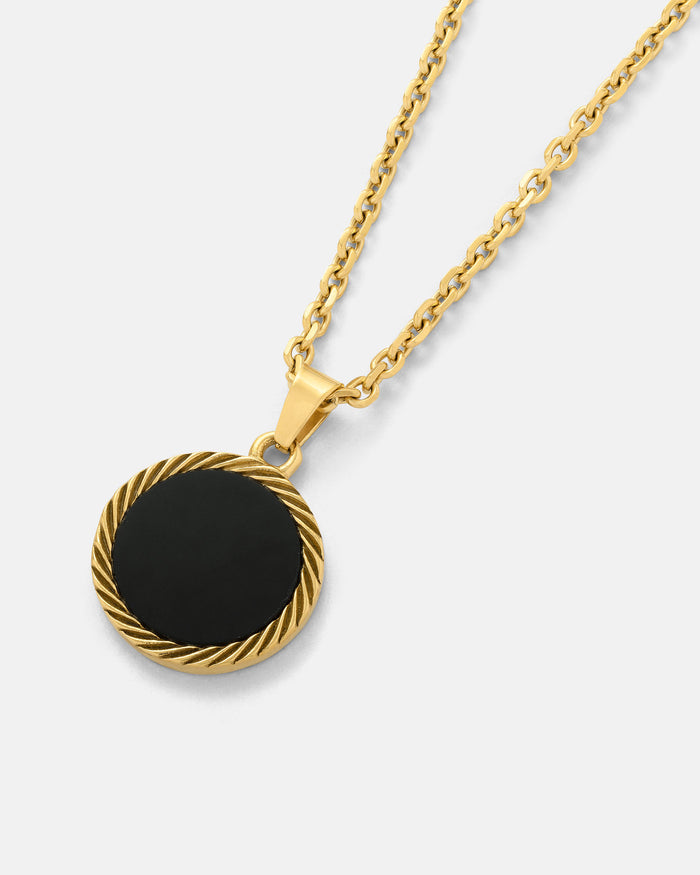 The Onyx Medallion Pendant (Gold) features a smooth black agate onyx set in a twisted gold rope border, elegantly displayed on a gold chain against a plain, light-colored background.
