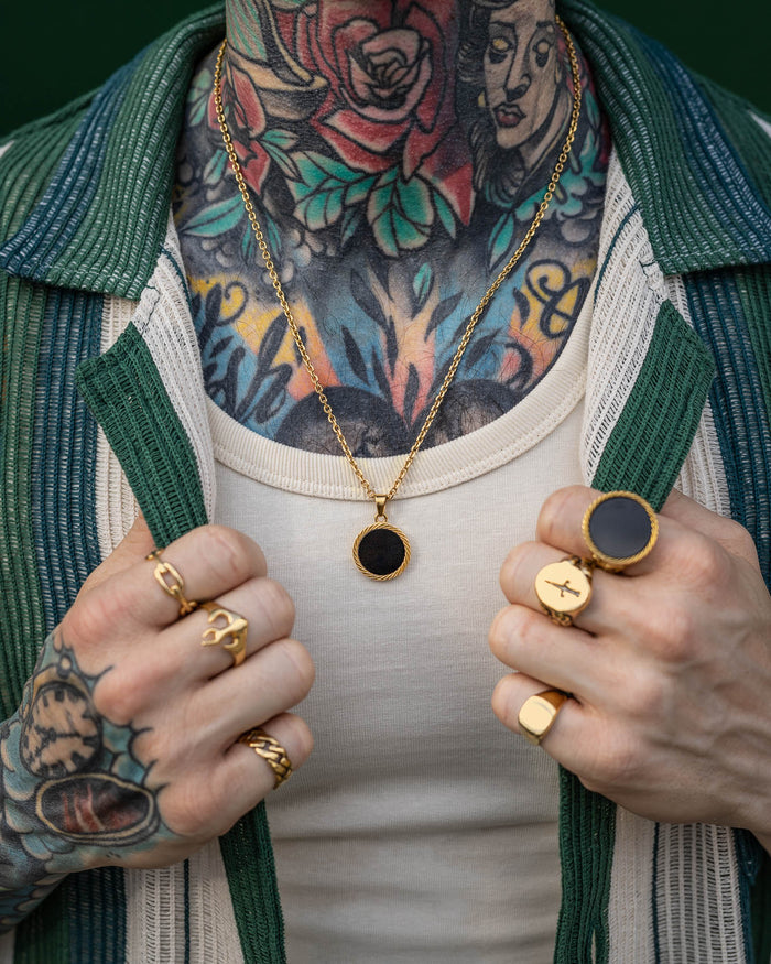 Wearing gold rings and the Onyx Medallion Pendant (Gold), a person with colorful chest and neck tattoos holds open a green and white striped shirt over a cream tank top.