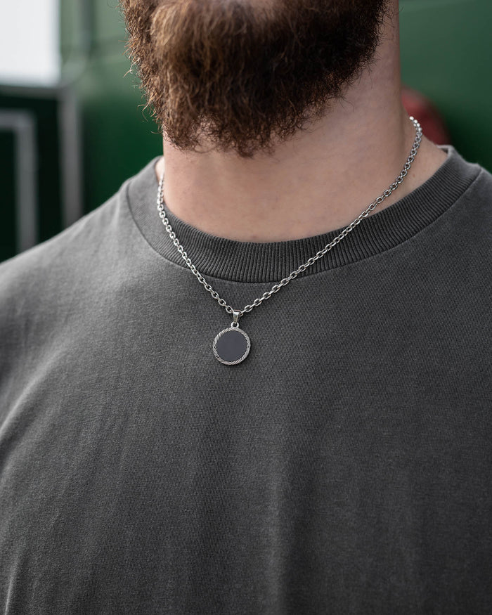A close-up of a bearded individual in a grey t-shirt models the Onyx Medallion Pendant on a silver chain, with a softly blurred green background.