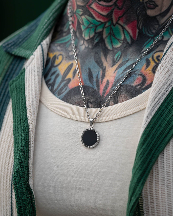 A close-up shows a person wearing the Onyx Medallion Pendant, featuring a round black charm. Their chest displays colorful floral and leaf tattoos, and they wear a cream shirt under an open, green ribbed shirt.