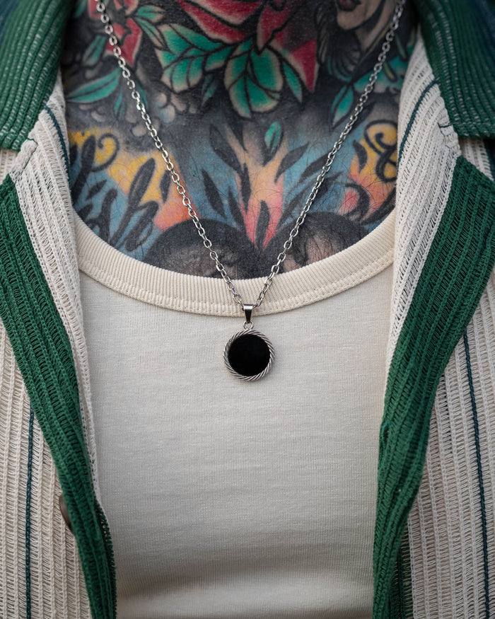 A close-up of a person wearing the Onyx Medallion Pendant over a cream-colored shirt, showing colorful floral neck tattoos and a green and white jacket.
