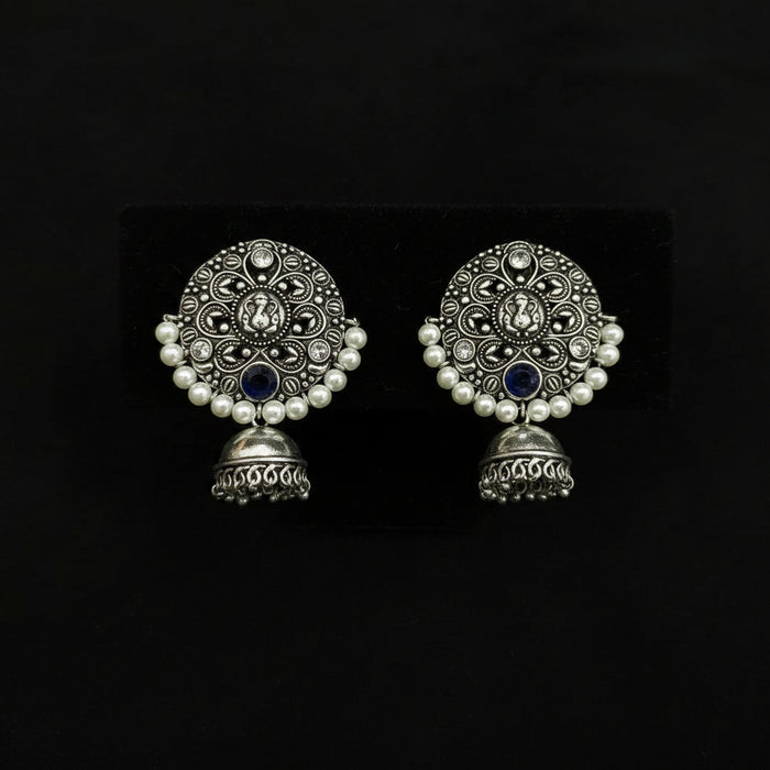 A pair of traditional Indian silver-toned jhumka earrings with intricate filigree work, white faux pearl borders, and dark blue stone accents, displayed on a black textured background.